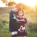 girl's left hand wrap around toddler while reading book during golden hour